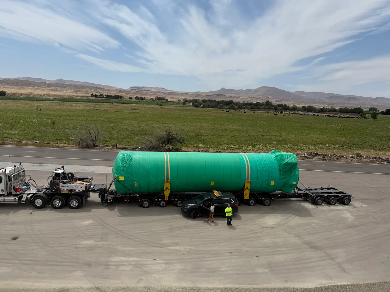 Full view of the long green-wrapped load, trailer and escort team at a roadside staging point